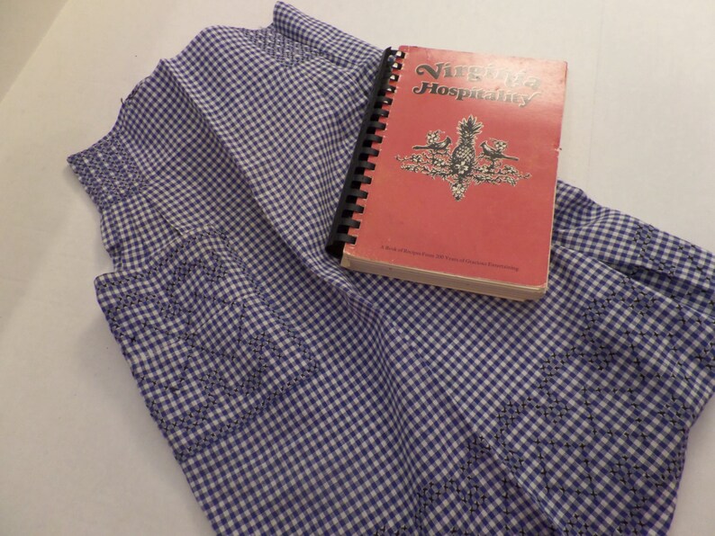 May include: A blue and white gingham fabric apron with a book titled "Virginia Hospitality" on top. The book has a red cover with a pineapple design.