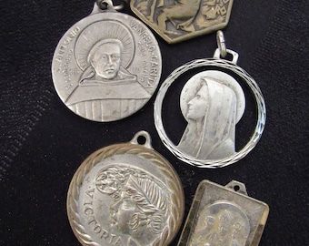 May include: Five silver and gold religious medallions with different images and text. The medallions feature images of a man with a beard, a woman with a crown, a woman with a veil, a man with a crown, and a man with a crown and a woman with a veil. The text on the medallions includes "S. ANTONIO", "VANGELO - CARITA", "VICTORIA", and "CRISTO RE".