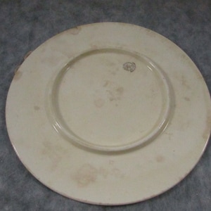 May include: Off-white ceramic plate with a raised center and a wide rim. The plate has a slightly aged appearance with subtle discoloration and a small emblem near the center. The plate is likely vintage or antique.
