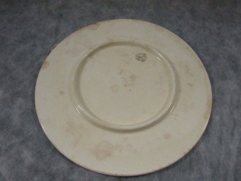 May include: Off-white ceramic plate with a raised center and a wide rim. The plate has a slightly aged appearance with subtle discoloration and a small emblem near the center. The plate is likely vintage or antique.