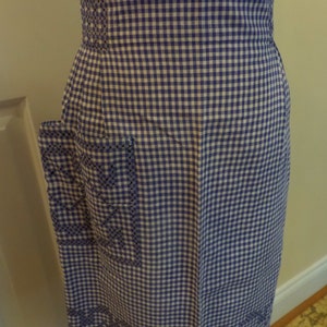 May include: A purple and white gingham apron with embroidered details. The apron has a large pocket on the left side and a waistband with a decorative stitch.