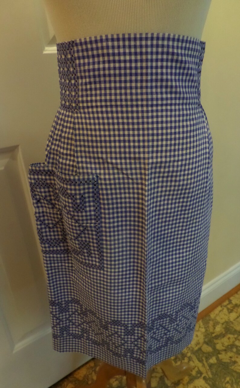 May include: A purple and white gingham apron with embroidered details. The apron has a large pocket on the left side and a waistband with a decorative stitch.