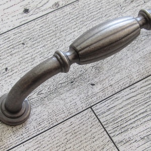 May include: A decorative cabinet pull with a curved handle and a central, ribbed design. The hardware is a muted, antique silver colour, and is designed for easy installation. The pull is approximately 15 cm long.