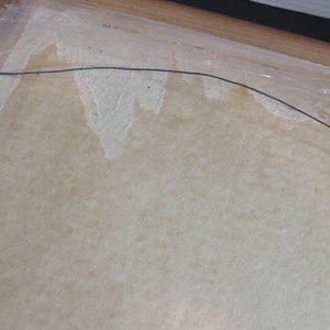 May include: A close-up of the back of a framed picture. The frame is made of wood and has a light brown finish. The picture is covered with a clear plastic sheet. A wire hanger is attached to the back of the frame.