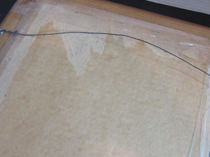 May include: A close-up of the back of a framed picture. The frame is made of wood and has a light brown finish. The picture is covered with a clear plastic sheet. A wire hanger is attached to the back of the frame.