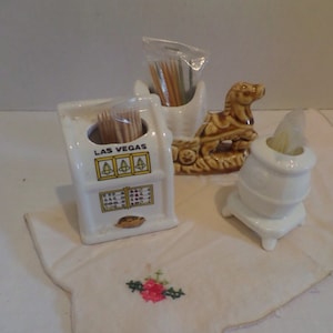 May include: Three ceramic toothpick holders. One is shaped like a slot machine with the words "LAS VEGAS" on it. Another is shaped like a horse-drawn carriage. The third is shaped like a wood-burning stove.