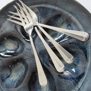 May include: A set of four stainless steel forks with slender handles and four tines, arranged in a fan pattern on top of a dark blue and black glazed ceramic oyster plate featuring six circular indentations for serving seafood.