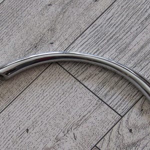 May include: A curved, polished chrome cabinet handle. The handle is a smooth, arc shape with mounting holes at each end. The shiny metal reflects light, creating highlights and shadows. This hardware is designed for cabinets, drawers, or other furniture.