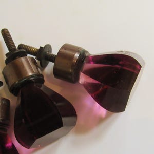 May include: Three cabinet knobs featuring faceted, translucent purple glass heads attached to dark metallic bases. Each knob includes a threaded metal bolt with a small circular washer and nut, designed for installation on furniture or doors.