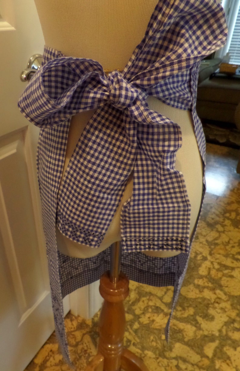May include: A blue and white gingham apron with a large bow tied at the waist. The apron is on a dress form.
