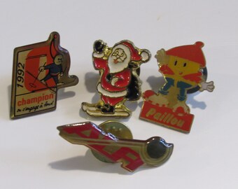May include: Four enamel pins featuring winter sports themes. One pin features a skier with the text "1992 champion". Another pin features a cartoon Santa Claus skiing. A third pin features a cartoon character skiing with the text "Pailllou". The fourth pin features a red and gold ski with the text "ALP".