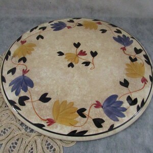 May include: A round, cream-colored decorative plate with a floral design. The plate features a black border and is adorned with yellow, blue, and red flowers with black leaves. The plate is approximately 10 inches in diameter.