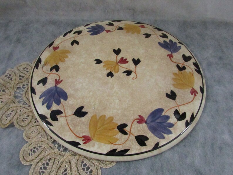 May include: A round, cream-colored decorative plate with a floral design. The plate features a black border and is adorned with yellow, blue, and red flowers with black leaves. The plate is approximately 10 inches in diameter.