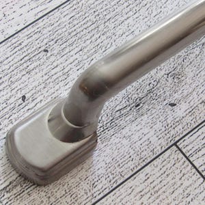 May include: A close-up of a brushed metal handle with a curved design. The handle has a rectangular base with a rounded top and a smooth, polished finish. The handle is likely for a cabinet or drawer.