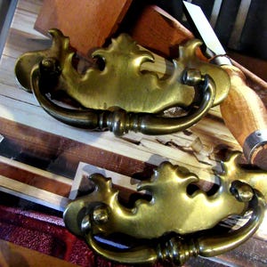 May include: Two matching brass drawer pulls featuring an ornate, curved backplate with a central decorative crest and a swinging bail handle. The metal has a polished, aged finish with visible highlights and shadows across the curved surfaces.