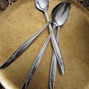 Puede incluir: Cuatro cucharas plateadas con detalles florales en los mangos dispuestas sobre un plato decorativo dorado. Las cucharas son reflectantes y el plato tiene una superficie texturizada. Las cucharas son probablemente para servir o comer.
