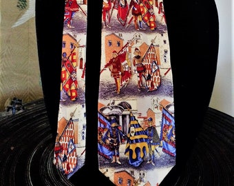 May include: A silk tie featuring a detailed print of a medieval parade scene with soldiers, flags, and buildings. The tie has a cream background with red, blue, and gold accents. The tie is displayed on a black mannequin.