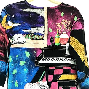 May include: Black long-sleeved shirt with a colorful print of a piano, a vase of flowers, a glass of wine, and two cats. The print is in a painterly style with a black background.