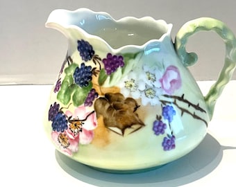May include: A porcelain creamer pitcher with a scalloped rim and a curved handle, featuring a hand-painted botanical design of dark purple blackberries, green leaves, thorny brown stems, and light pink and white flowers on a pale green base.