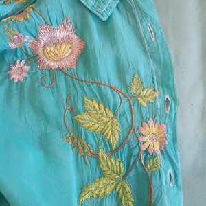 May include: A turquoise button-down shirt featuring intricate embroidery of pink flowers, yellow centers, and green leaves. The fabric has a crinkled texture, and the design includes a pointed collar with matching floral stitching along the neckline.
