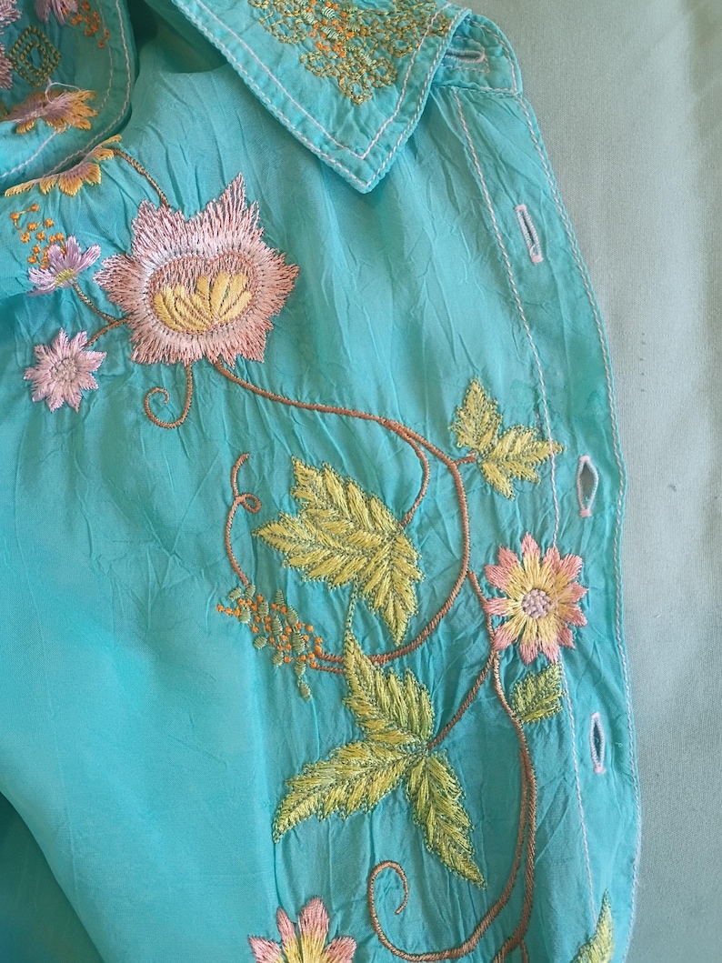 May include: A turquoise button-down shirt featuring intricate embroidery of pink flowers, yellow centers, and green leaves. The fabric has a crinkled texture, and the design includes a pointed collar with matching floral stitching along the neckline.