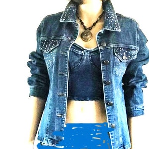 May include: A dark blue denim jacket featuring silver stud embellishments on the collar and chest pockets, worn over a dark blue denim crop top. The jacket has a button-front closure and long sleeves, paired with a beaded necklace with a round pendant.