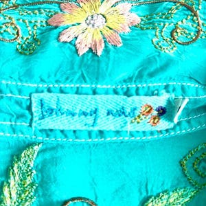 May include: A bright turquoise shirt featuring intricate embroidery of a multi-colored flower with pink and yellow petals, surrounded by swirling brown vines and green leaves. A rectangular fabric label reads Johnny Was in blue thread.