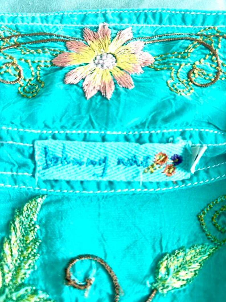 May include: A bright turquoise shirt featuring intricate embroidery of a multi-colored flower with pink and yellow petals, surrounded by swirling brown vines and green leaves. A rectangular fabric label reads Johnny Was in blue thread.