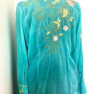 May include: A long-sleeved turquoise button-down shirt featuring intricate floral embroidery in shades of pink, yellow, and brown. The design includes detailed leaf patterns and decorative stitching across the back and on the wide cuffs.