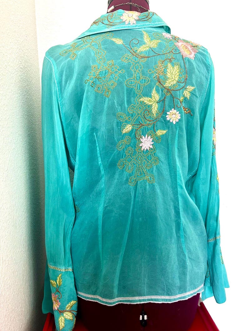 May include: A long-sleeved turquoise button-down shirt featuring intricate floral embroidery in shades of pink, yellow, and brown. The design includes detailed leaf patterns and decorative stitching across the back and on the wide cuffs.