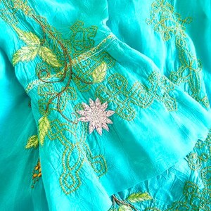 May include: A bright turquoise garment featuring intricate lime green embroidery in geometric and leaf patterns. A single pale pink flower applique is centered on the flared sleeve, accented by thin brown vine-like stitching details.