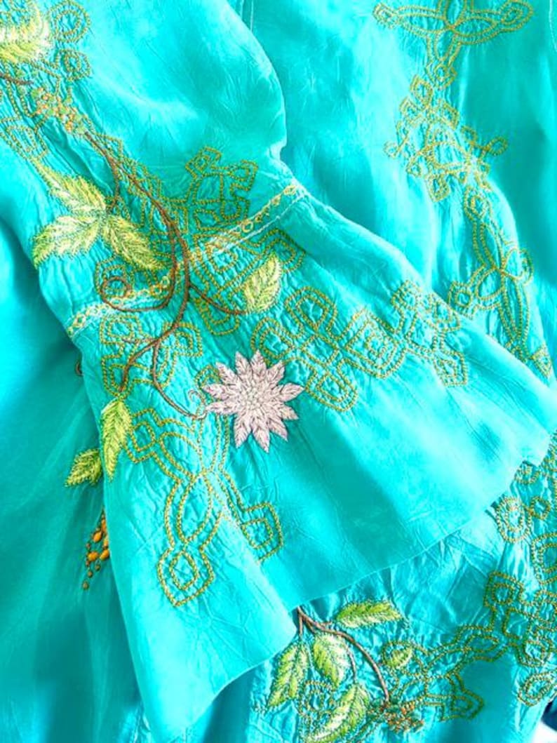 May include: A bright turquoise garment featuring intricate lime green embroidery in geometric and leaf patterns. A single pale pink flower applique is centered on the flared sleeve, accented by thin brown vine-like stitching details.