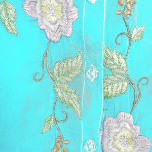 May include: A bright turquoise shirt featuring intricate floral embroidery in shades of silver, white, and pale green. The garment includes a vertical button placket with clear, round buttons and white stitching along the center seam.