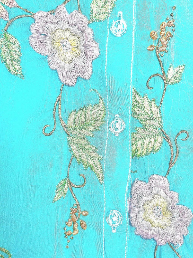 May include: A bright turquoise shirt featuring intricate floral embroidery in shades of silver, white, and pale green. The garment includes a vertical button placket with clear, round buttons and white stitching along the center seam.