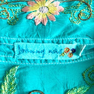 May include: A bright turquoise fabric garment featuring intricate embroidery of a central pink and yellow flower with green stems. A rectangular fabric label is stitched below the flower, displaying the text Johnny Was in blue thread.