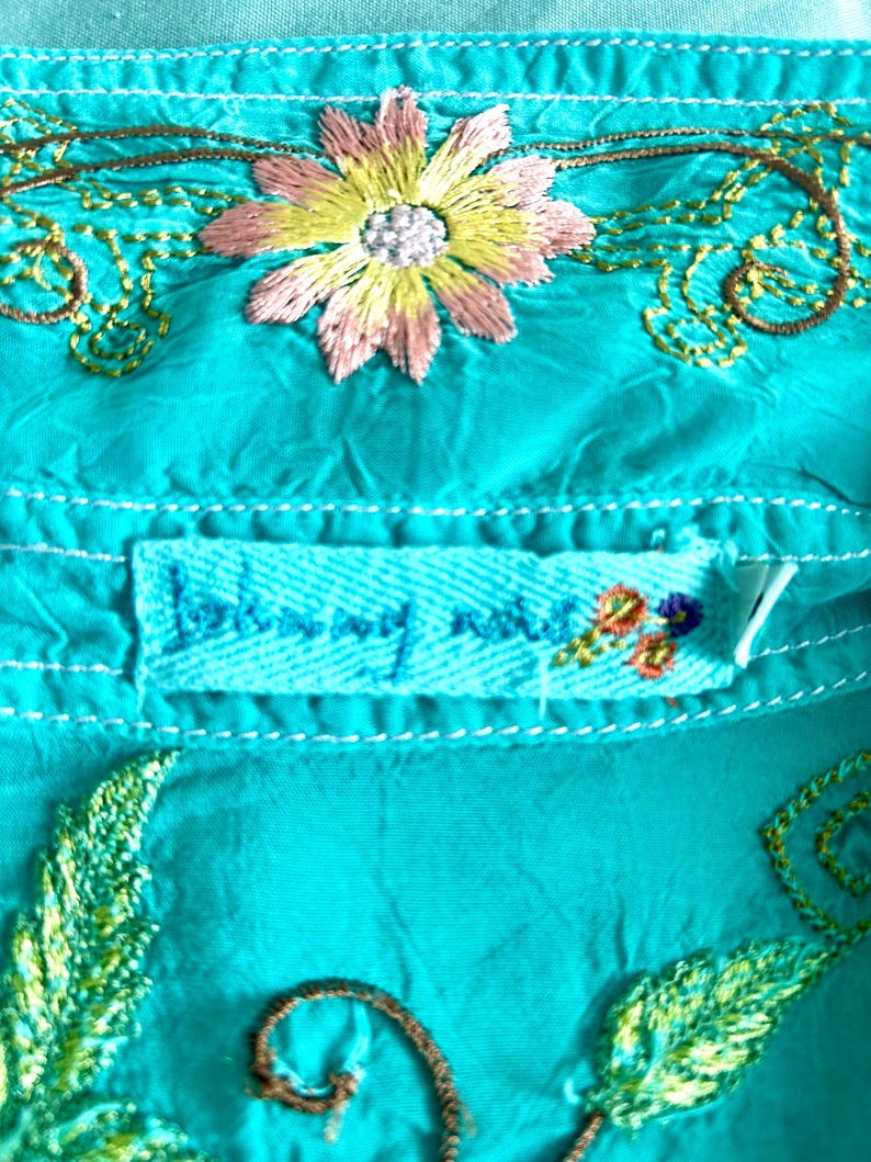 May include: A bright turquoise fabric garment featuring intricate embroidery of a central pink and yellow flower with green stems. A rectangular fabric label is stitched below the flower, displaying the text Johnny Was in blue thread.