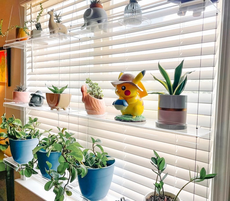 May include: A set of clear acrylic shelves with various plants and figurines. The shelves are mounted on a window with white blinds. The figurines include a yellow Pikachu, a white rabbit, and a white elephant. The plants are in small pots and include succulents, ferns, and a snake plant.