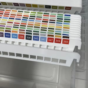 May include: A close-up of a white organizer drawer filled with color swatches. The swatches are arranged in rows and columns, displaying a variety of colors. Each swatch is labeled with a number. The drawer is partially open.