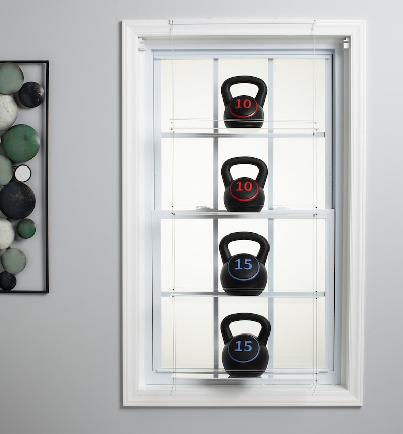 May include: Four black kettlebells hanging on a clear shelf system inside a white window frame. The kettlebells are labeled with the numbers 10, 10, 15, and 15.
