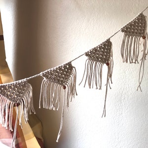May include: A macrame banner with triangular flags and wooden beads. The banner is made of natural beige cotton cord and has a rustic, bohemian style.