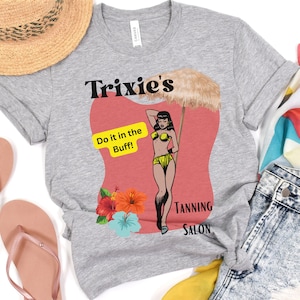 May include: A gray t-shirt with a retro-style illustration of a woman in a bikini under a beach umbrella. The text on the shirt reads "Trixie's Do it in the Buff! Tanning Salon".