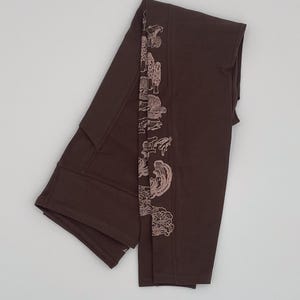 May include: Dark brown leggings featuring a vertical row of light pink line art illustrations of various mushrooms along the side. The leggings have a high waistband with visible seam detailing and a smooth, matte fabric construction throughout.