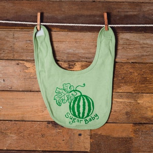 May include: A green baby bib with a watermelon and leaf design and the text "Sugar Baby".