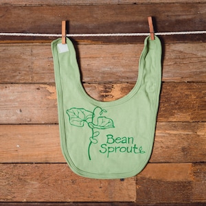 May include: A light green baby bib with a green bean sprout design and the words "Bean Sprout" printed on it. The bib is hanging from a clothesline with wooden clothespins against a wooden background.
