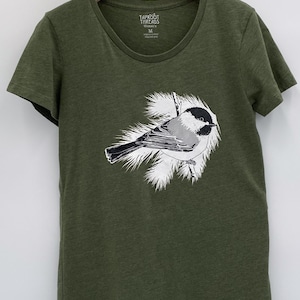 Puede incluir: Camiseta verde oliva con cuello redondo, que muestra una ilustración detallada de un pájaro. El pájaro tiene plumas negras, blancas y grises y está posado en una rama con follaje blanco. El nombre de la marca "TAPROOT THREADS" está impreso en la etiqueta.