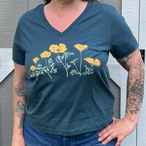Puede incluir: Una camiseta azul turquesa con cuello en V y un estampado gr&aacute;fico de amapolas amarillas de California.