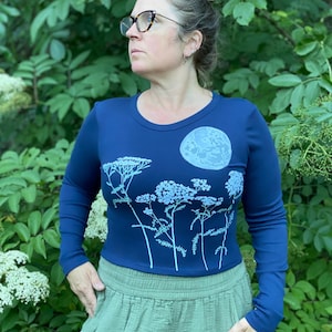 May include: A navy blue long-sleeved shirt with a round neck featuring a light blue floral design and a moon graphic. The shirt is paired with olive green pants. The background is filled with green foliage.