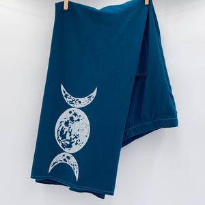 May include: A teal-colored pajama set hanging from a clothesline. The top features a white graphic of three moon phases. The pajama set is made of soft material and is hanging with wooden clothespins.
