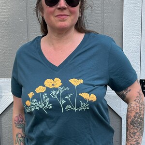 Puede incluir: Una camiseta azul turquesa de cuello en V con un estampado gr&aacute;fico de amapolas amarillas de California. La camiseta tiene mangas cortas y un corte holgado.