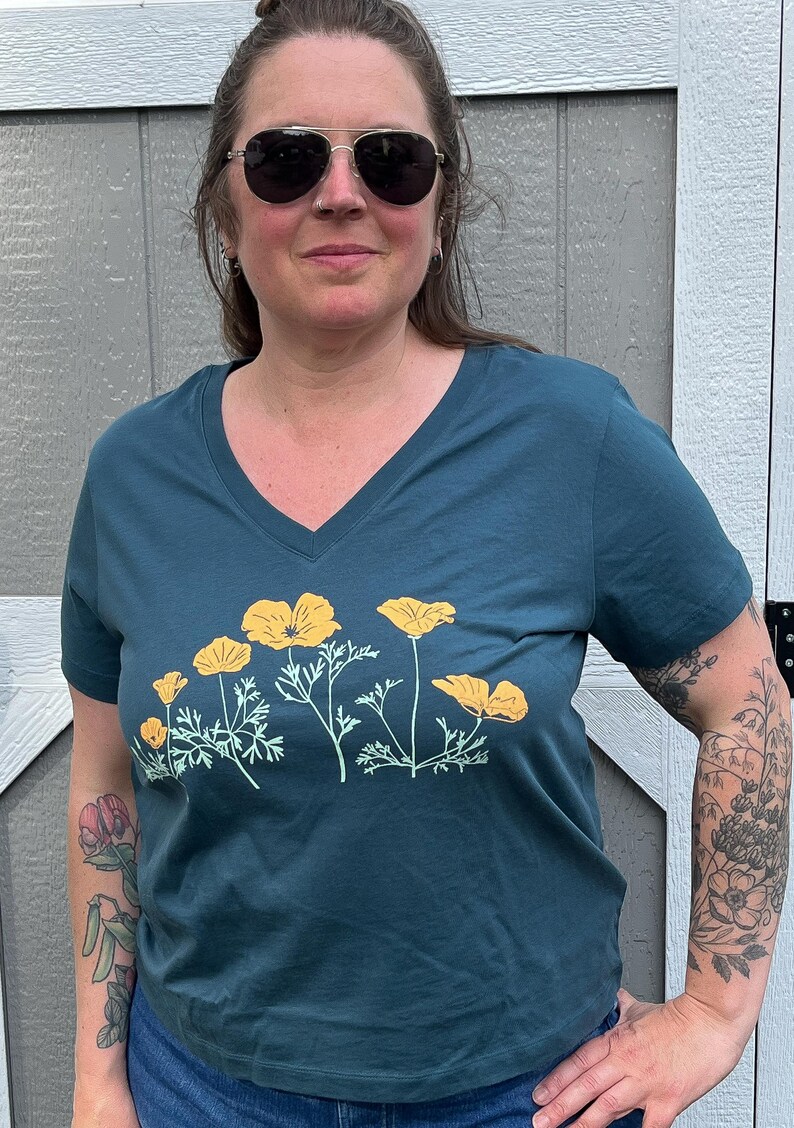 Puede incluir: Una camiseta azul turquesa de cuello en V con un estampado gr&aacute;fico de amapolas amarillas de California. La camiseta tiene mangas cortas y un corte holgado.